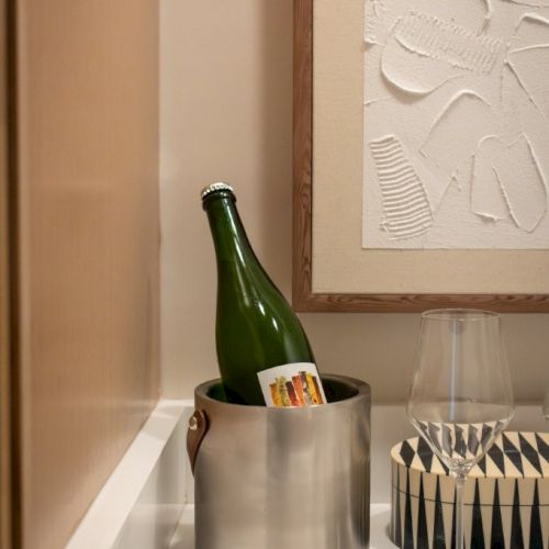 A bottle in an ice bucket, a wine glass, and a coaster on a white counter beside framed artwork.