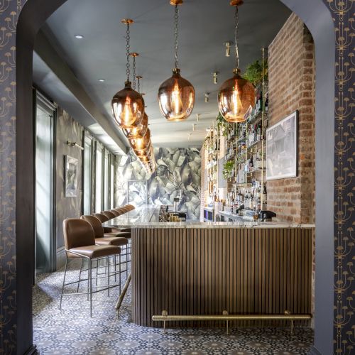A stylish bar features pendant lights, patterned flooring, and a wooden counter with seating, set in an elegant arched alcove.