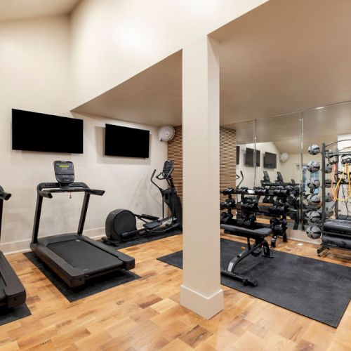 A modern gym with treadmills, mirrors, exercise bikes, free weights, and stability balls on a wooden floor, equipped with large screens.