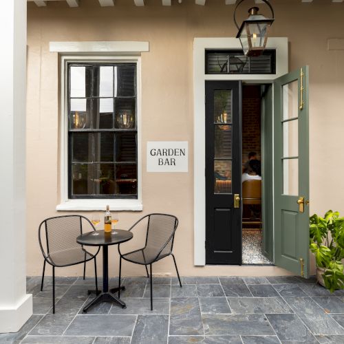 Outdoor seating area with round tables, chairs, a bottle, and a "Garden Bar" sign on a wall beside open doors leading to an indoor space.