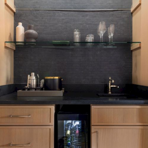 This image shows a small home bar with wooden cabinets, a glass shelf holding glassware, and a countertop with a sink and drinkware.