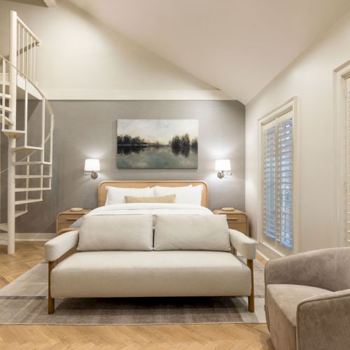 A modern bedroom with a spiral staircase, bed, armchair, and wall art, featuring white shutters and parquet flooring, creating a cozy ambiance.