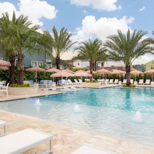 A luxurious pool area with loungers, palm trees, and striped umbrellas under a blue sky with fluffy clouds.