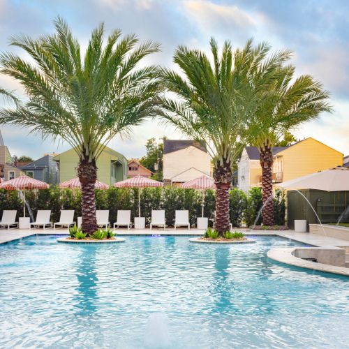 A tranquil pool scene with palm trees, sun loungers, and colorful houses in the background under a clear blue sky.