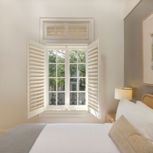 A cozy bedroom with an open window, shutters, a bed with white linens, a bedside lamp, and a framed artwork on the wall.