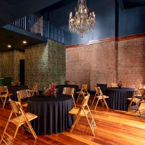 The image shows a room with wooden floors, round tables with black cloths, wooden chairs, brick walls, and a chandelier.