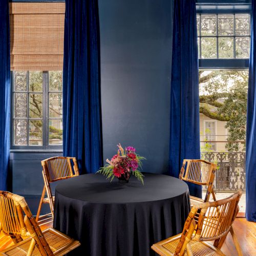 A room with dark blue walls, a round table with a black tablecloth and flowers, surrounded by four wooden chairs, and two large windows.