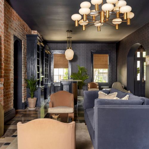 A stylish living room with a brick wall, navy furniture, a modern chandelier, and large windows, creating a cozy, sophisticated ambiance.