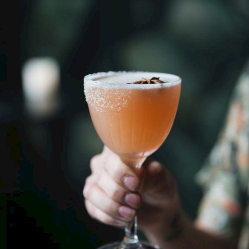 A hand is holding a cocktail glass with a frothy, orange drink, garnished with star anise, against a dark, blurred background.