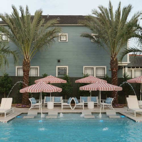 A swimming pool with lounge chairs and striped umbrellas, set against a building and palm trees in the background.