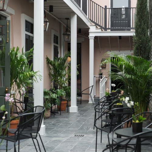 Outdoor patio with black metal chairs, tables, and lush green plants along a tiled path under a balcony.