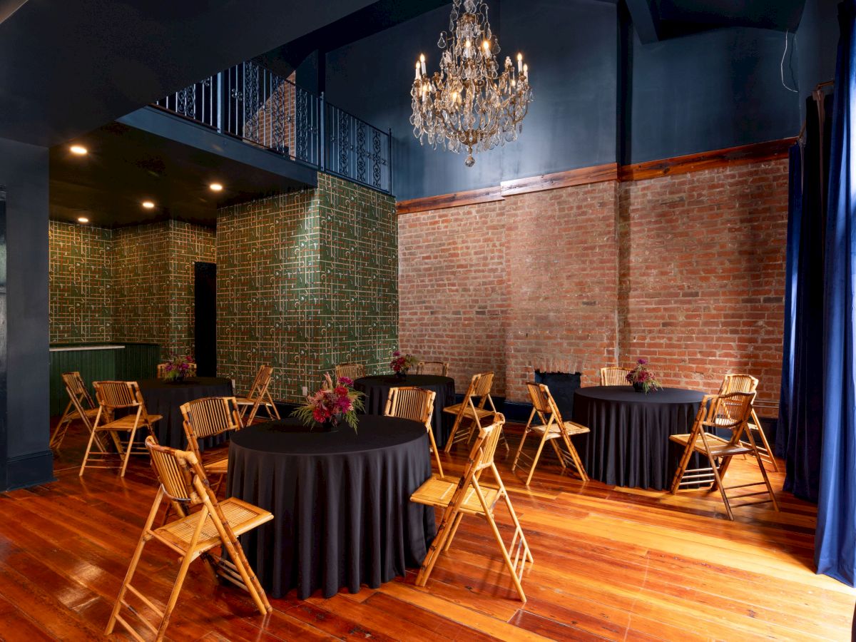 The image shows a room with wooden flooring, round tables with black tablecloths, wooden chairs, exposed brick walls, and a chandelier.
