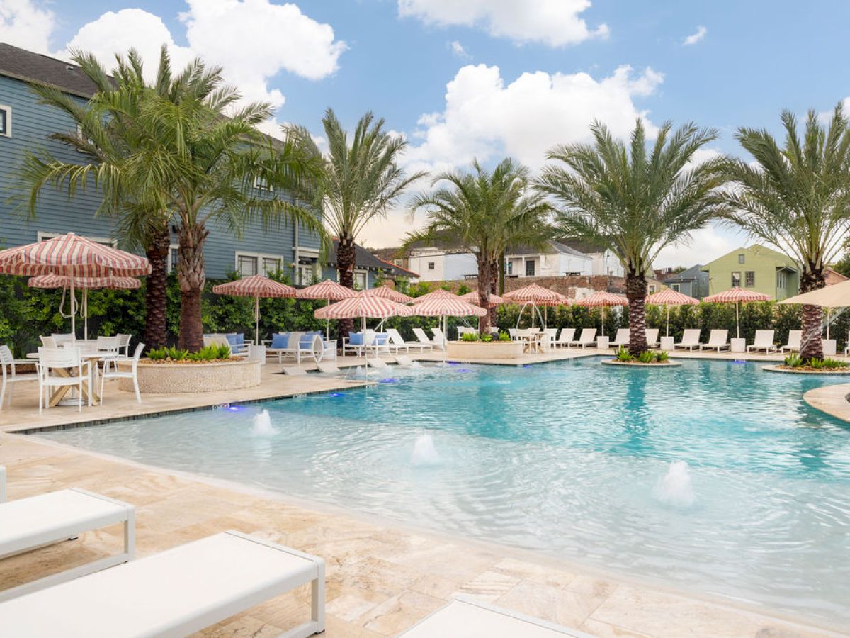 A swimming pool surrounded by palm trees, sun loungers, and striped umbrellas, under a partly cloudy sky.