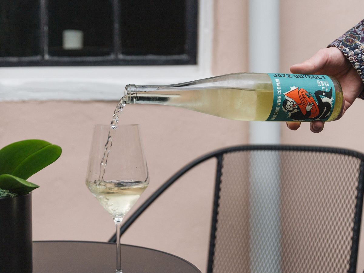 A hand pours white wine from a bottle into a glass on a black metal table with a chair and a small plant nearby.