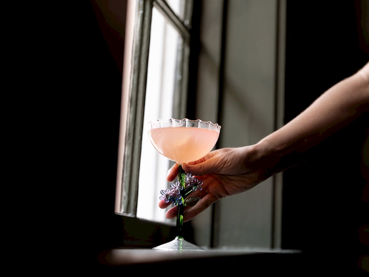 A hand holding a pink beverage in an elegant glass, near a window with dark surroundings.