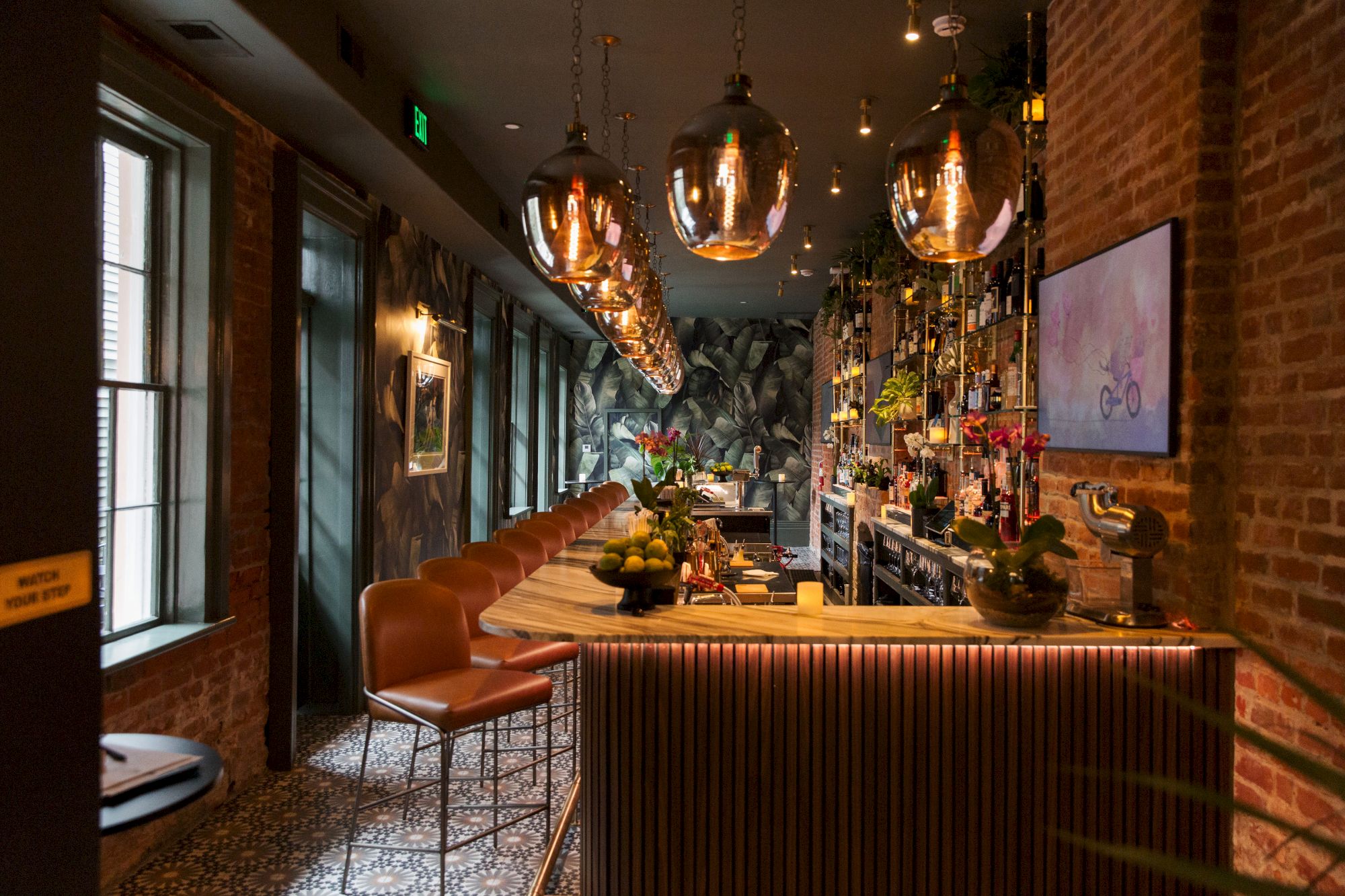 A stylish bar with leather stools, hanging lights, and a brick wall featuring a TV and artwork creates a cozy atmosphere.