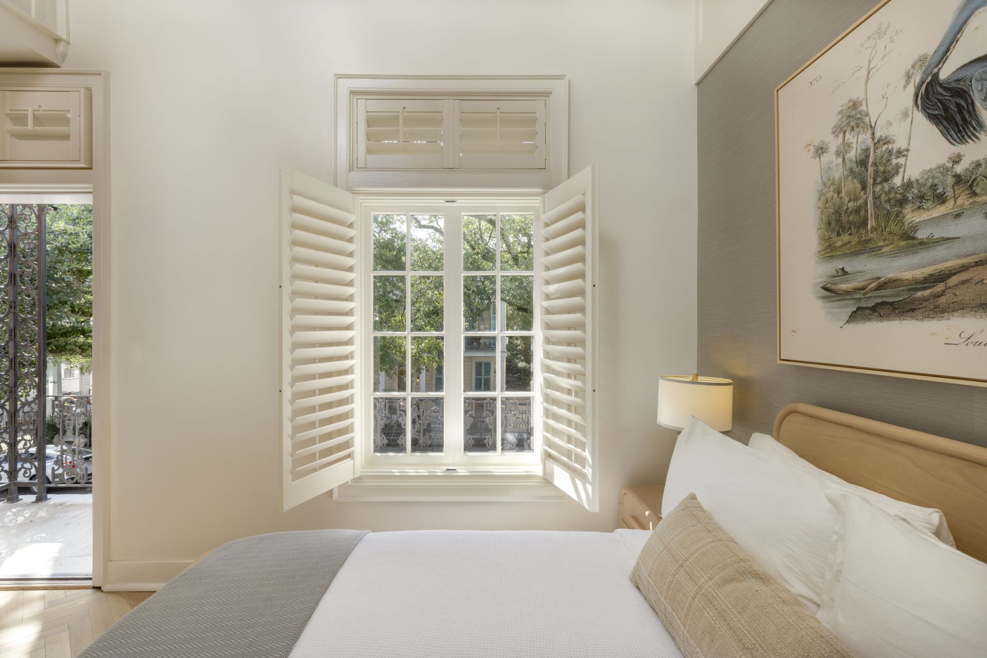 A cozy bedroom with a bed, open window shutters letting in light, artwork on the wall, and a lamp on a small table.