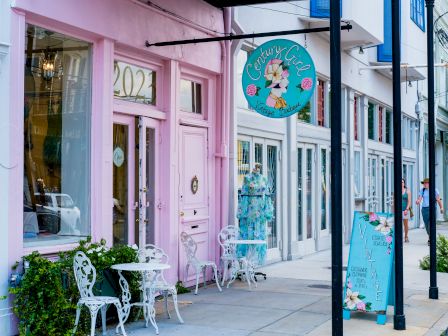 A pastel pink storefront with a decorative round sign, outdoor seating, and a teal sandwich board on the sidewalk.