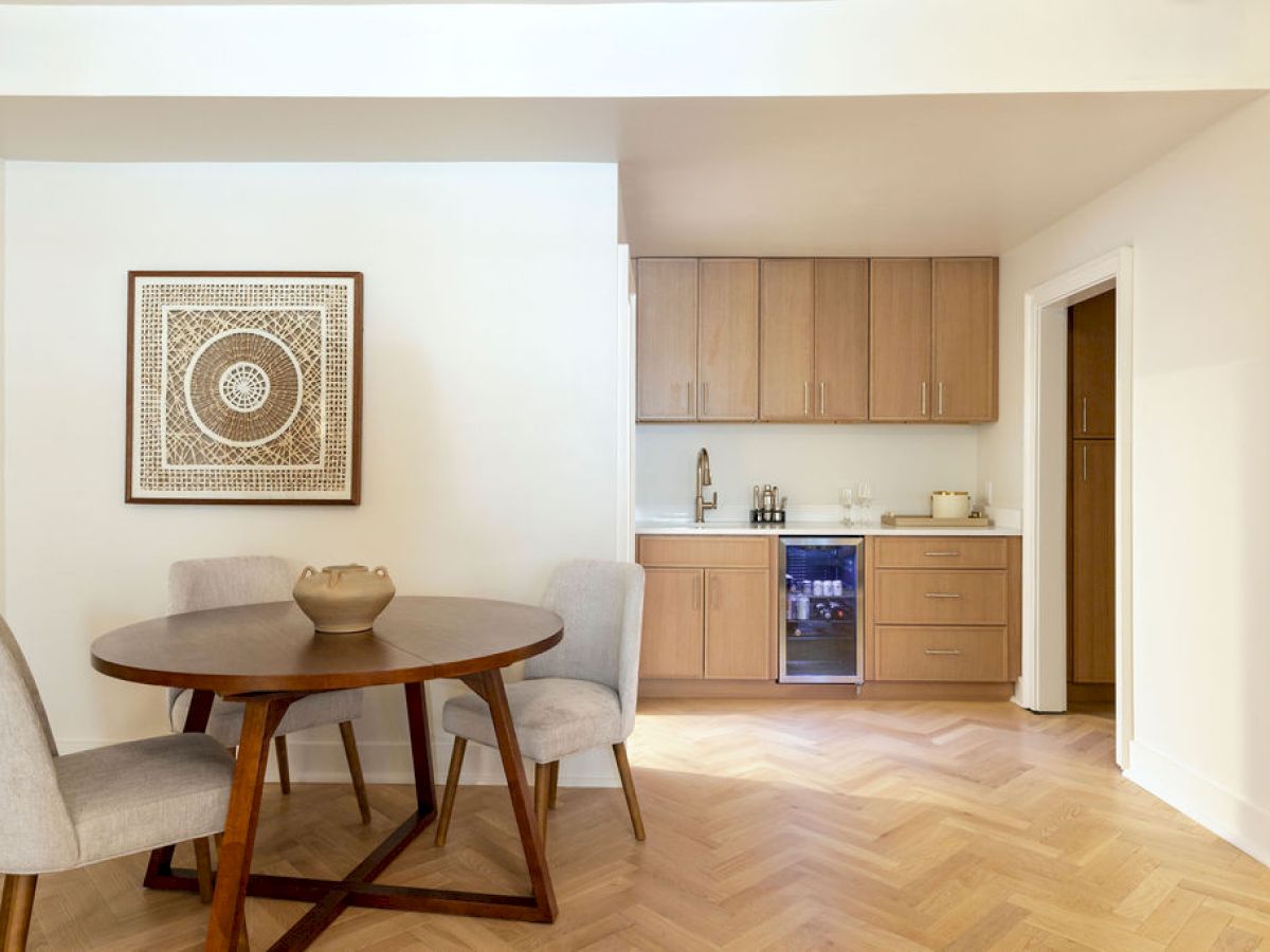The image shows a cozy dining area with a round table, four chairs, artwork on the wall, and a small kitchenette with wooden cabinets and appliances.