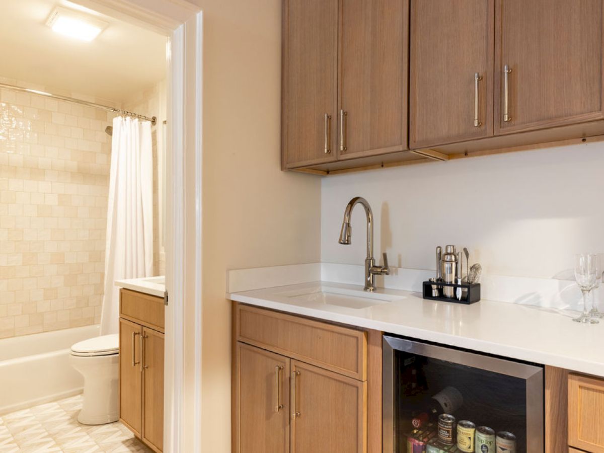 The image shows a bathroom and kitchenette with wood cabinets, a sink, a small fridge, and various kitchen items.