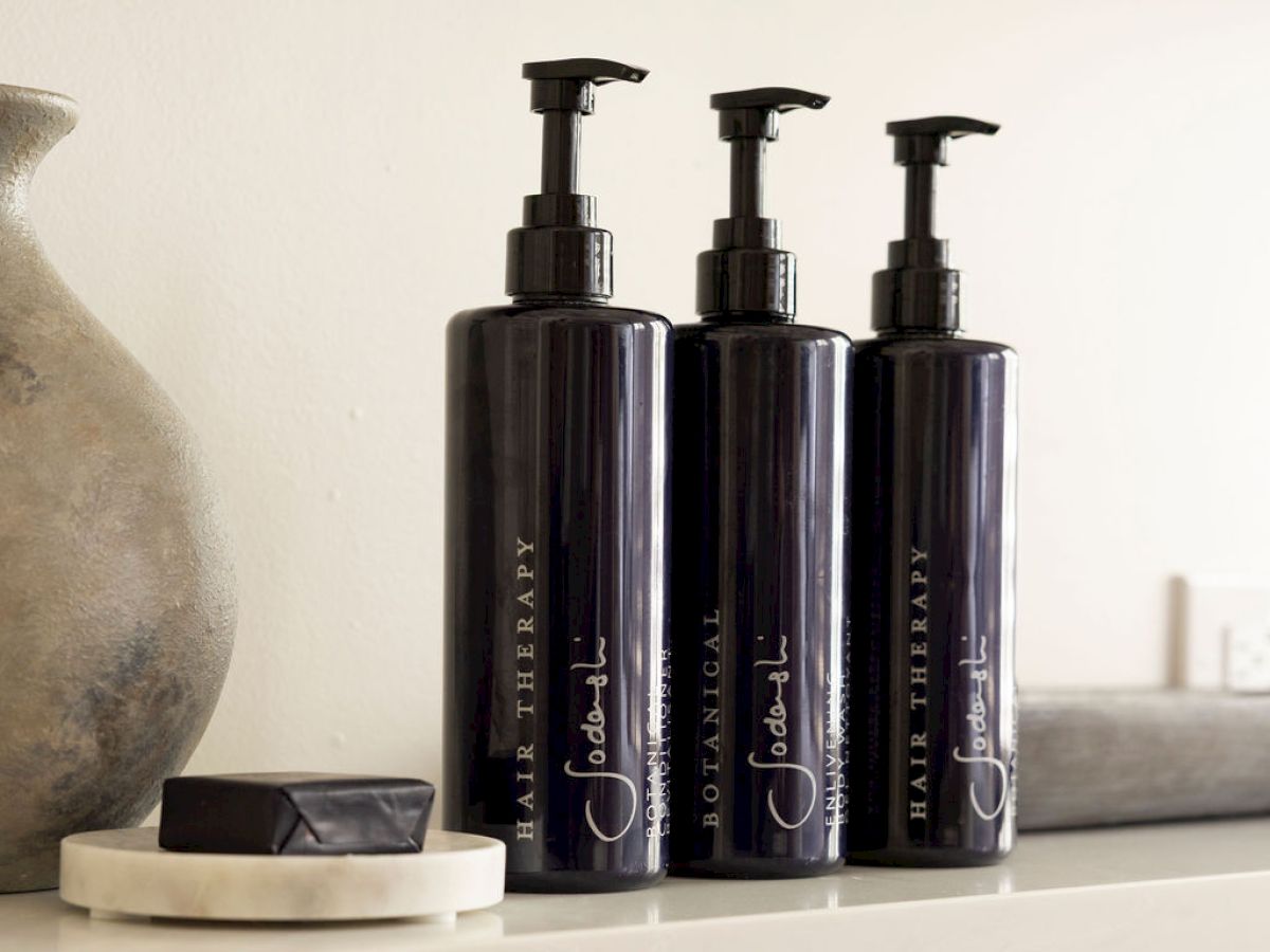 The image shows three dark pump bottles labeled "Hair Therapy" on a shelf, accompanied by a ceramic piece and a small dish with a dark item.