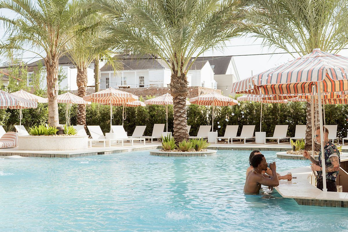 A sunny pool area with palm trees, people seated at a swim-up bar, and loungers under striped umbrellas can be seen in the image.