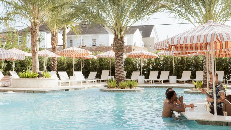 A sunny pool area with palm trees, people seated at a swim-up bar, and loungers under striped umbrellas can be seen in the image.