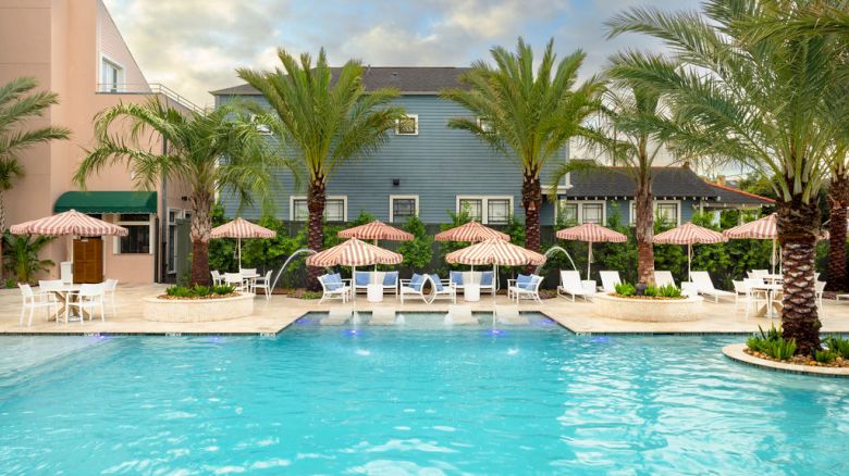 A serene pool surrounded by lounge chairs, umbrellas, palm trees, and adjacent buildings, creating a relaxing resort-like atmosphere.