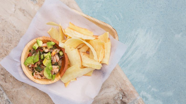 A bowl of ceviche with avocado sits next to plantain chips on a tray by a pool.