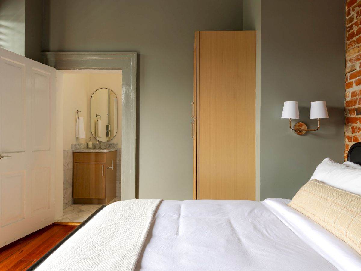 A cozy bedroom with a neatly made bed, brick wall accent, and a view into an adjacent bathroom featuring a modern sink and round mirror.