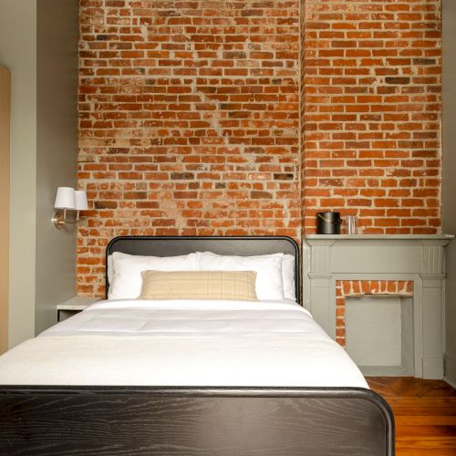 A bedroom with exposed brick wall, wooden floor, bed with white bedding, light wood wardrobe, and a small mantle.