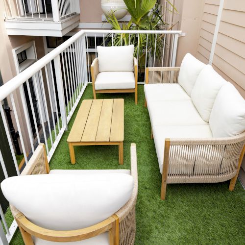 A small balcony with white cushioned seating and a wooden table, surrounded by white railings and artificial grass flooring.
