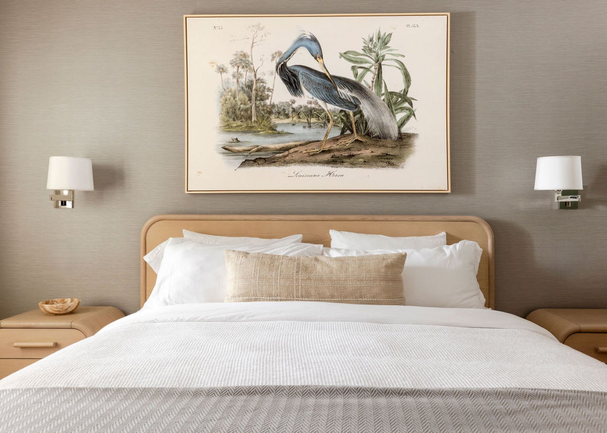 A cozy bedroom features a neatly made bed with white linens, flanked by wall-mounted lamps, and a framed artwork of birds above.