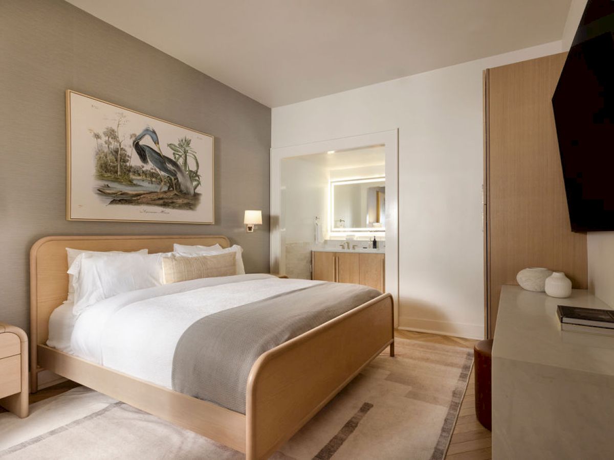 The image shows a modern hotel room with a large bed, a painting of a bird, a TV, and a bathroom visible through an open door.