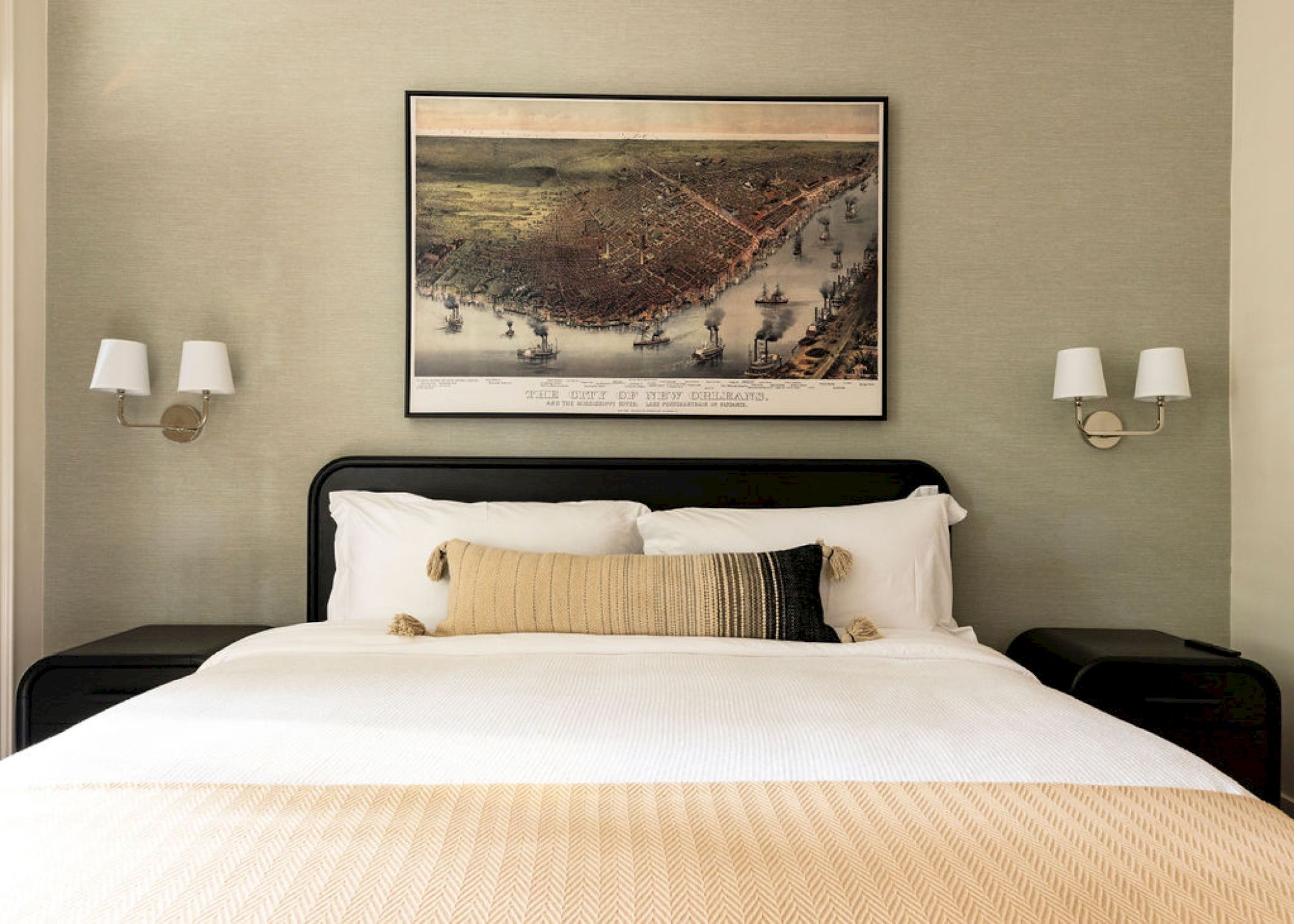 A neatly made bed with pillows, flanked by nightstands and wall-mounted lamps, is under a framed artwork depicting a historic coastal scene.