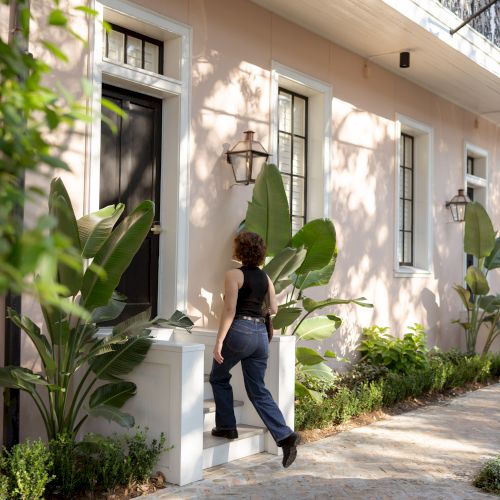 A person is walking toward the entrance of a pastel-colored building, surrounded by lush greenery and large potted plants.
