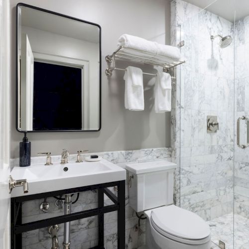A modern bathroom with marble walls, a glass shower, a rectangular mirror, and a white sink, with towels neatly placed on a rack.