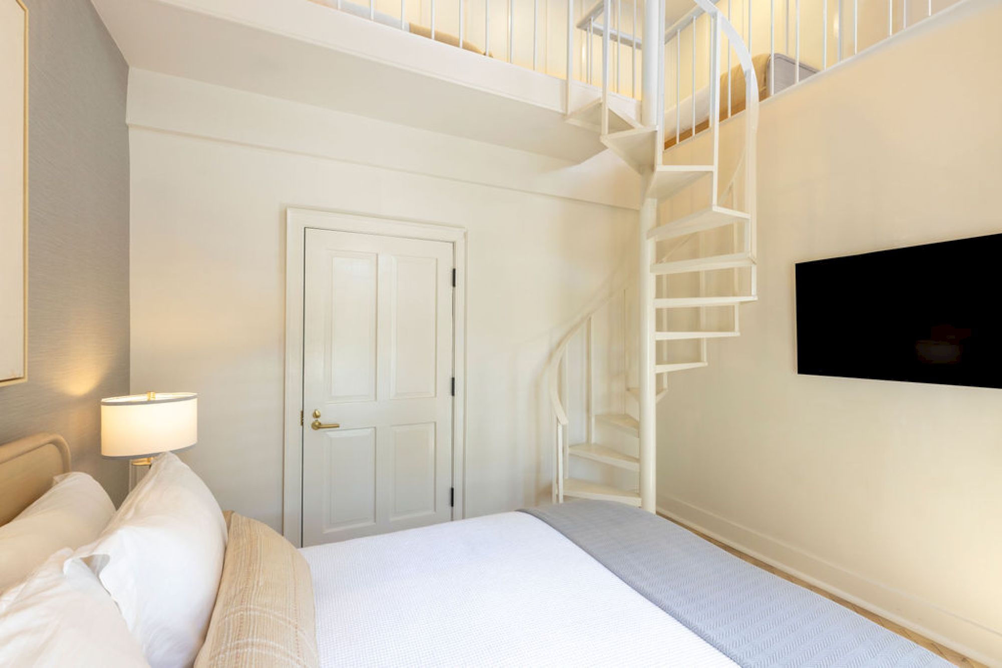 A cozy bedroom features a bed, a spiral staircase leading to a loft, a door, a wall-mounted TV, and a lamp, all in neutral tones.