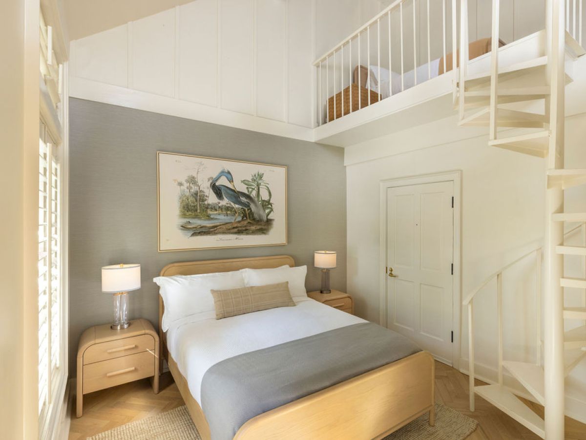 A cozy bedroom features a bed with a gray blanket, wooden furniture, spiral staircase leading to a loft, and a pelican artwork on the wall.