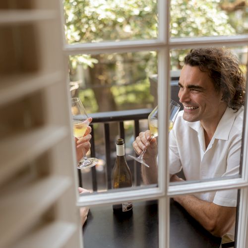 A person is sitting at an outdoor table, smiling with a drink in hand, seen through a window.