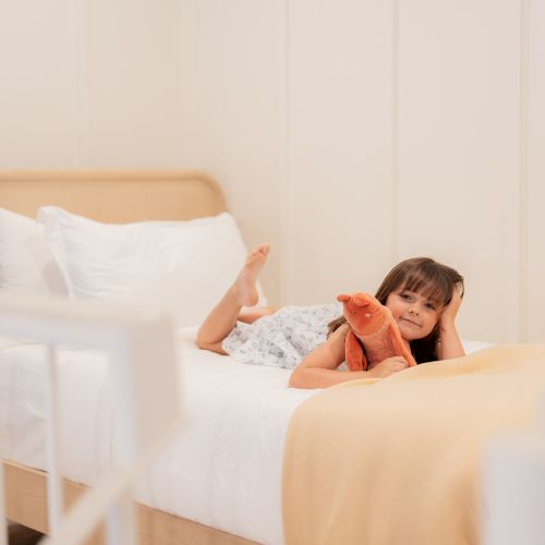 A child is lying on a bed with white sheets and a beige blanket, in a softly lit room with a warm and cozy ambiance.