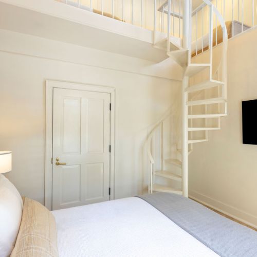 The image shows a modern bedroom with a bed, a spiral staircase, a door, a wall-mounted TV, and a nightstand with a lamp.