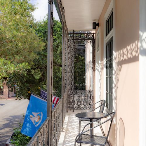 A balcony with ornate ironwork, a small table and chairs, and two flags overlooking a street lined with trees.