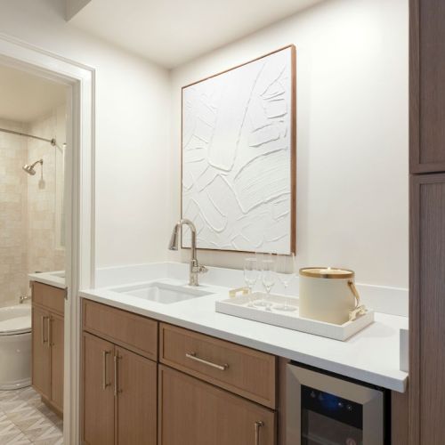 The image shows a kitchenette with wooden cabinets and a white countertop, adjacent to a bathroom. A textured artwork hangs above the sink.