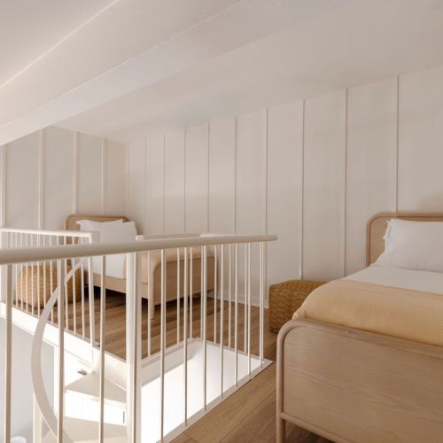 The image shows a loft bedroom with two single beds, wooden flooring, white walls, and a railing overlooking the floor below.