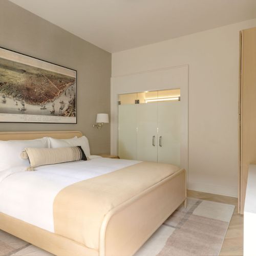 A modern bedroom with a double bed, beige color scheme, artwork on the wall, and minimalist decor, featuring a glass-door closet.