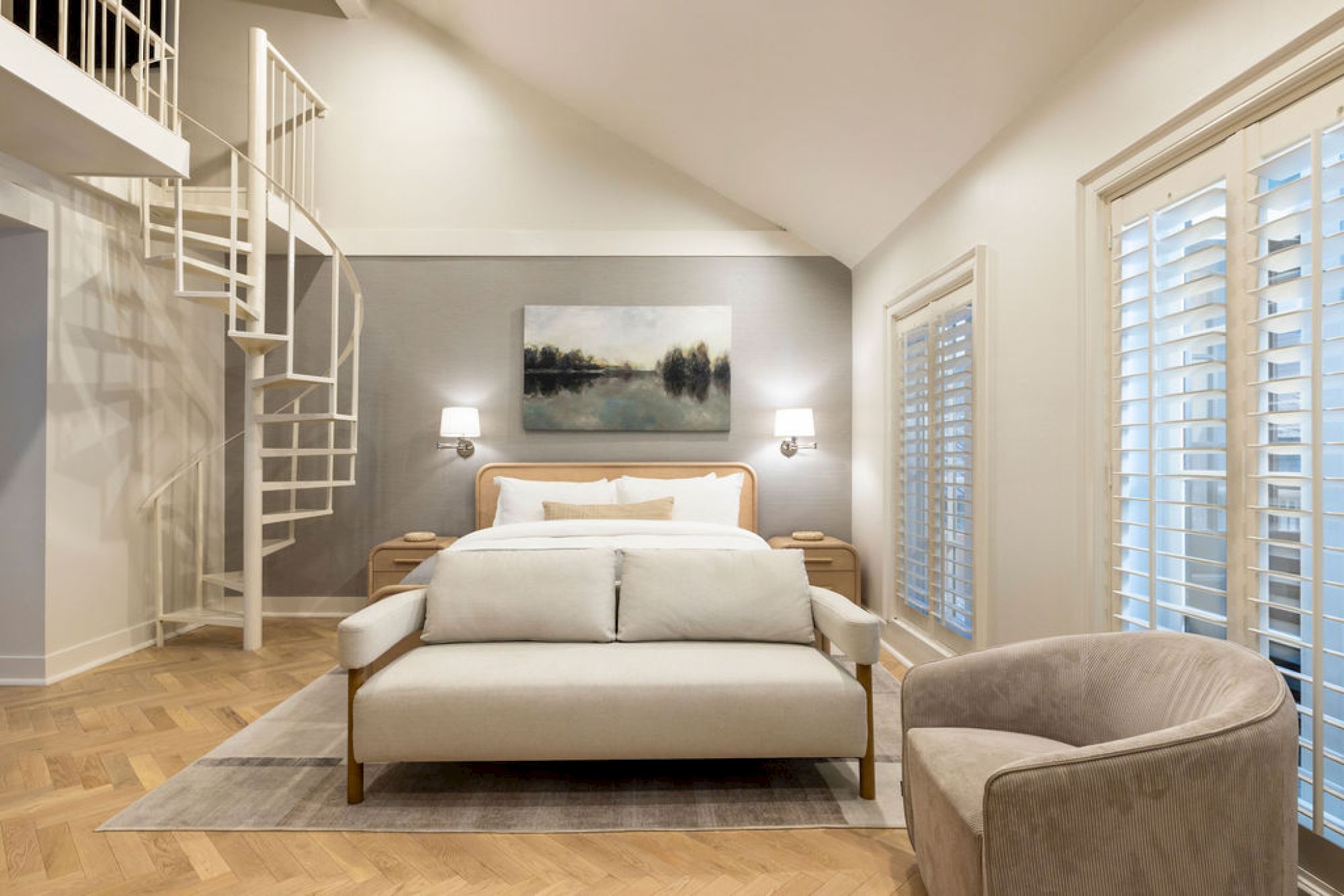 This image shows a modern bedroom with a spiral staircase, a large bed, artwork on the wall, and a comfy chair next to windows with shutters.