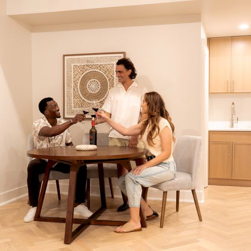 Three people are sitting around a table, toasting with glasses. There's a decorated wall and a kitchenette in the background.