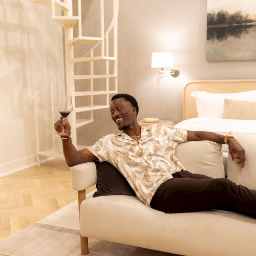 A person lounges on a couch holding a wine glass, near a spiral staircase in a cozy room with a bed and artwork.