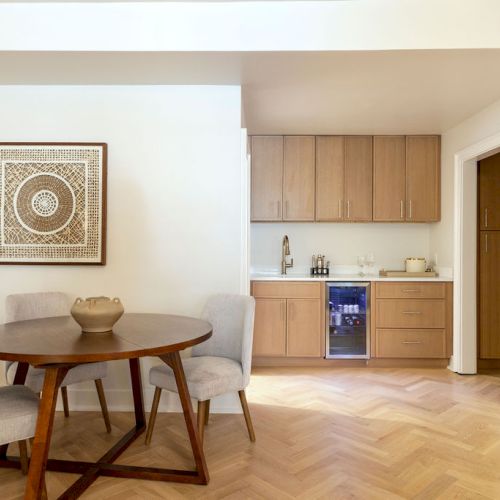 A dining area with a round table, three chairs, wall art, and a kitchenette with wooden cabinets and appliances.